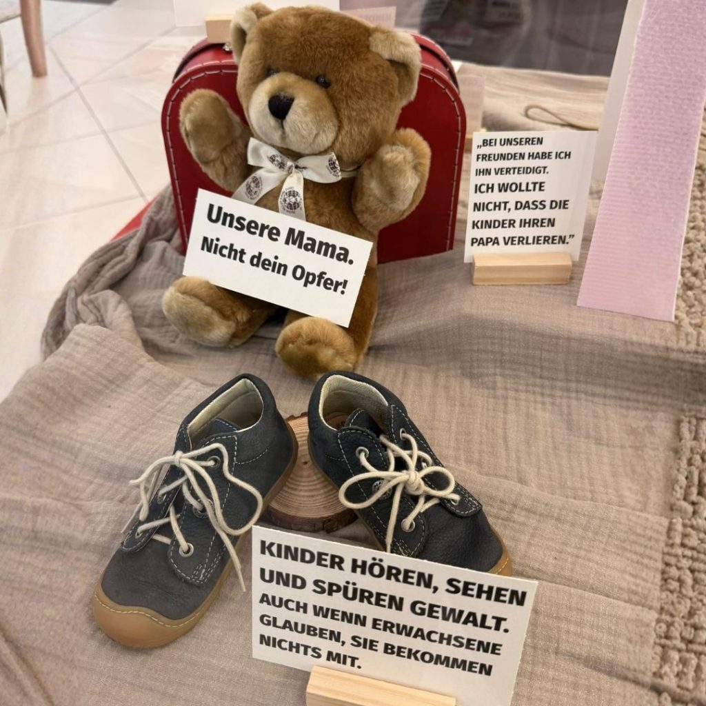 Ein Teddybär sitzt neben zwei Kinderschuhen auf einem Stoff. Es sind Schilder mit Botschaften gegen Gewalt in der Familie sichtbar.