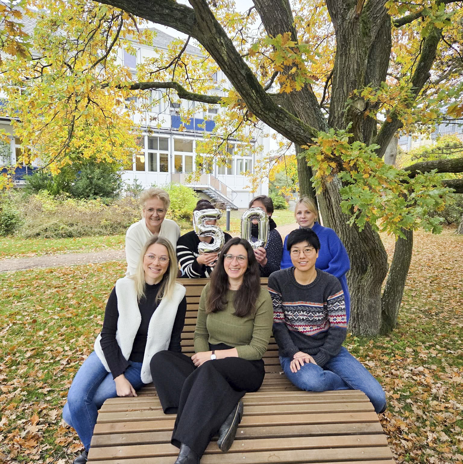 Eine Gruppe von Menschen sitzt auf einer Holzbank unter einem Baum im Freien.
