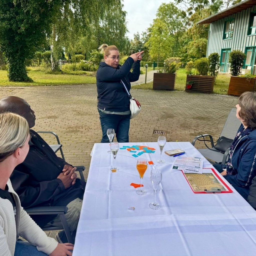 Menschen sitzen an einem Tisch im Freien, eine Person steht und spricht zu ihnen.