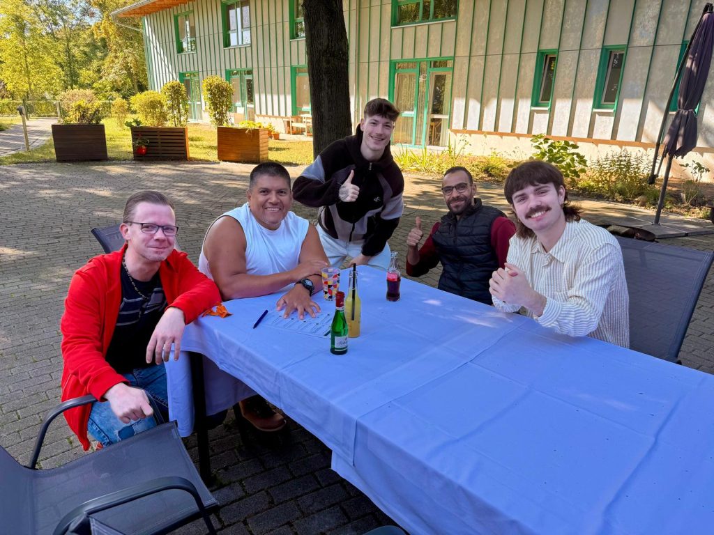 Eine Gruppe von fünf Männern sitzt an einem Tisch im Freien, umgeben von grüner Vegetation und einem Gebäude im Hintergrund.